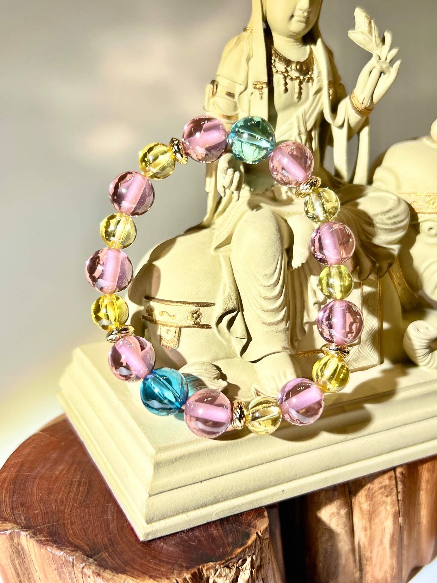 三色水龍珠手串由藍色、黃色與粉紅色晶珠組成，置於米白色雕像上，晶珠透亮而柔和，呈現細膩光感與溫暖氛圍